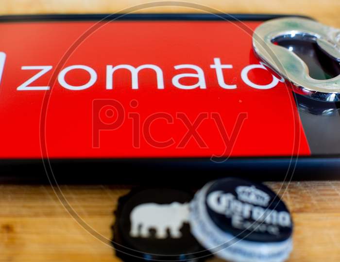 Mobile Phone Showing Zomato On A Wooden Platform With Beer Bottle Caps And An Opener On The Side