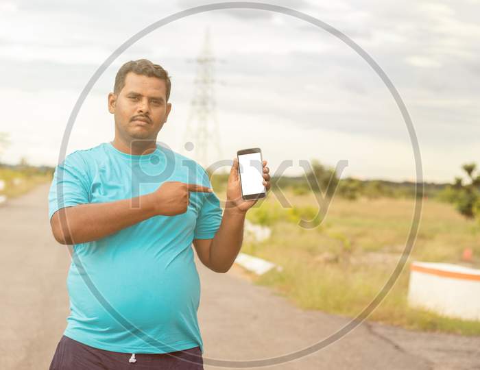 Young Man Wearing Jogging Dress Showing Smartphone Screen By Pointing With Hand And Finger At Park - Concept Of Recommended Fitness Tracker App On Mobile.