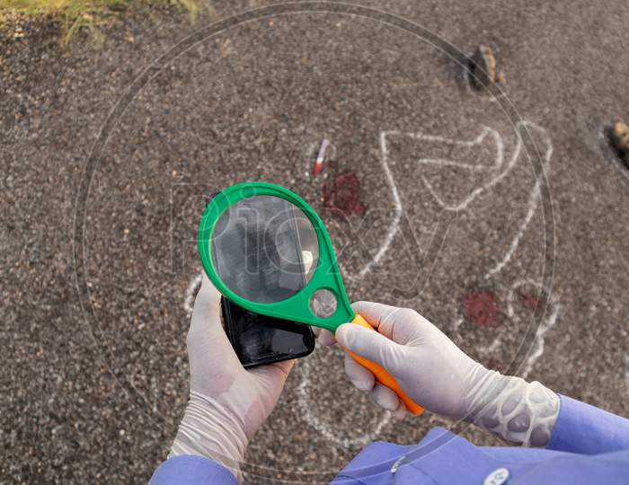 Detective Or Investigator Verifying The Fingerprints On Mobile Using Magnifier And Chalk Outline Dead Body As A Background