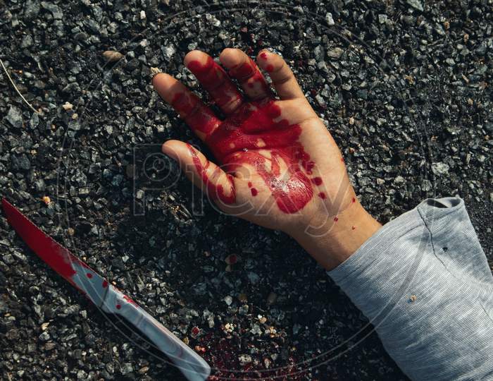 Close Up Of Hand With Blood And Bloody Knife On Road.