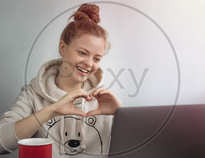Young Caucasian woman with laptop at home making a video call, talking and smiling, showing a heart shape gesture. Stay in touch with loved ones conce