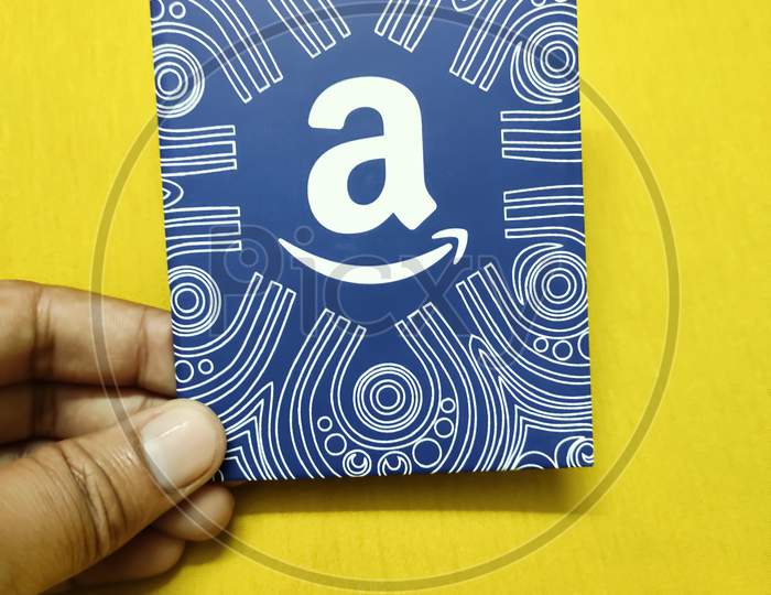 A hand holds a Amazon gift card against yellow background, which allows the recipient to purchase items from the Amazon.com website