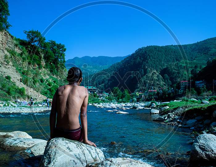 Young Boy Sitting On A Rock Near Flowing River Looking At The Beautiful Mountains Landscape