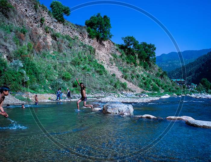 Himachal Pradesh, India - April 20Th, 2019: Playful Children Swimming In Fresh River Water On A Summer Vacations Surrounded By Beautiful Green Hill Mountains