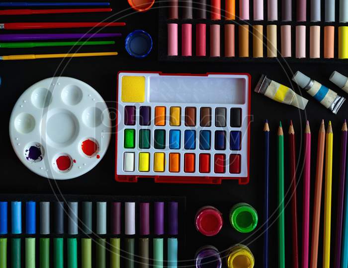 Colorful stationery flat lay: pastel crayons, colored pencils, watercolors on black background. Art workspace school concept.