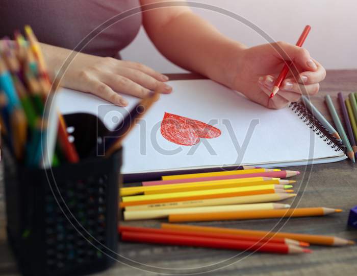 Close up of woman hands drawing a heart with colored pencils on white paper. Art studio workplace concept