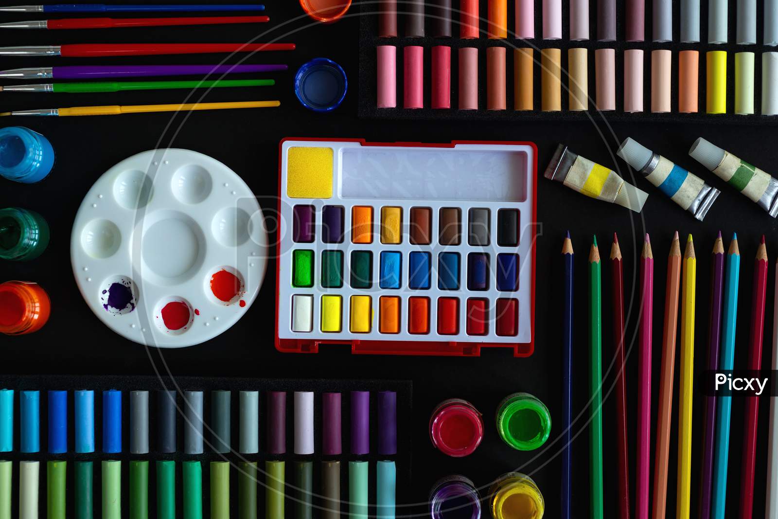 Colorful stationery flat lay: pastel crayons, colored pencils, watercolors on black background. Art workspace school concept.