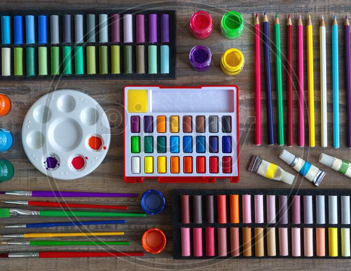Colorful stationery flat lay: pastel crayons, colored pencils, watercolors on wooden background. Art workspace school concept. Top view