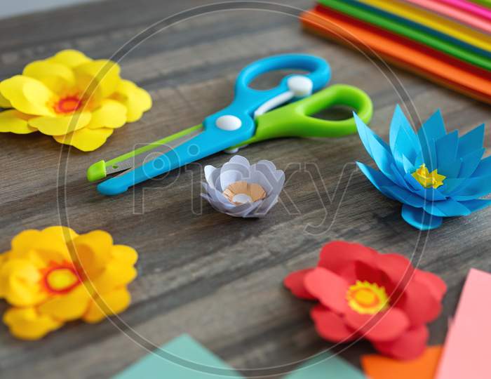 Close up of colorful color paper flowers and scissors on wooden desk. Art studio workplace concept