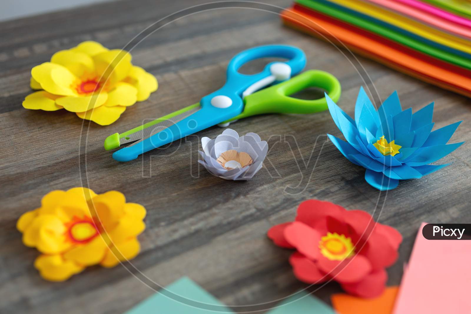 Close up of colorful color paper flowers and scissors on wooden desk. Art studio workplace concept