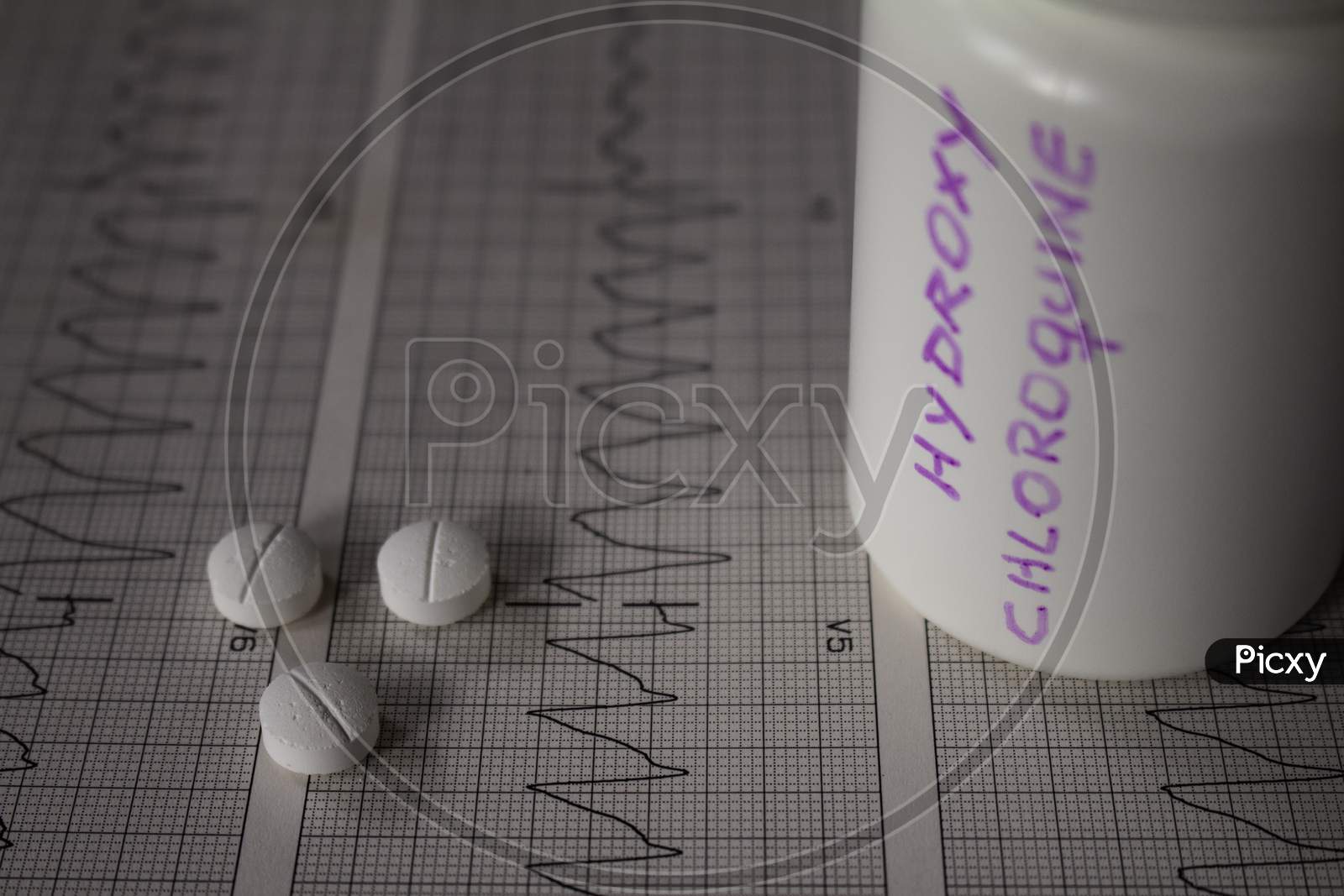 Adverse Effects On The Heart By Hydroxychloroquine Or Chloroquine. Pills On An Electrocardiogram With Cardiac Arrhythmias. White Container With "Hydroxychloroquine" Written On The Side.