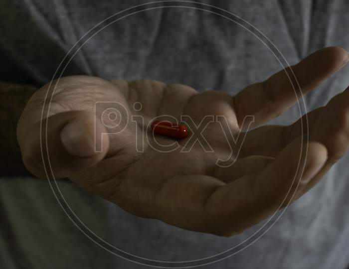 Hand Of A Man Wearing A Gray T-Shirt Holding A Red Capsule In His Hand.