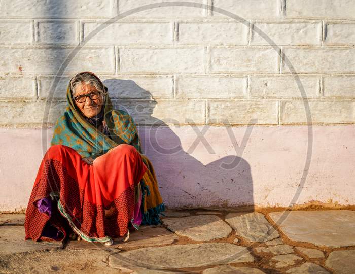 Old Aged Woman Sitting With The Support Of The Wall In The Sunset Wearing Glasses