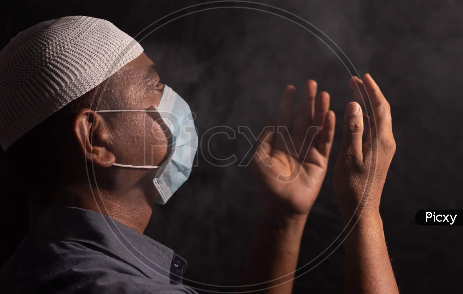 Muslim Man With Medical Mask And Cap Praying To God In Dark Room To Protect Or Save From Covid-19 Or Coronavirus Crisis - Spirituality And Surrender Concept.