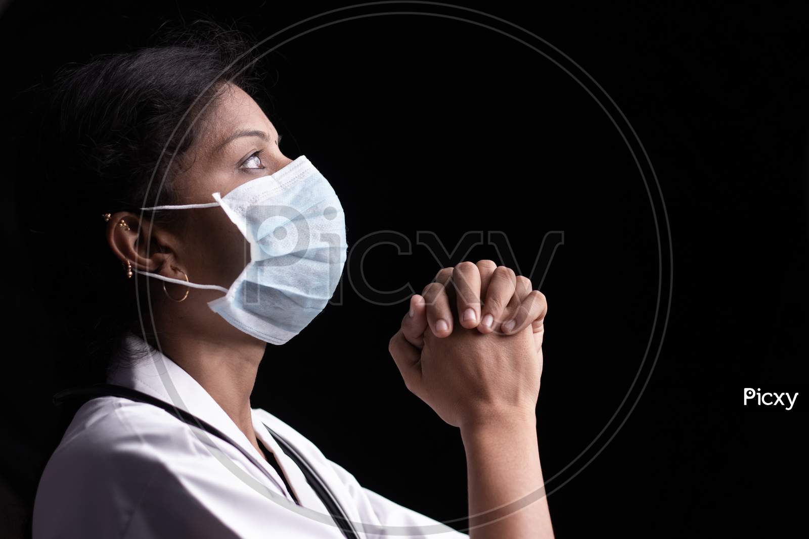 Profile View Of Young Woman Doctor In Medical Mask Praying To God On Black Background Looking Up - Concept Of Hope And Fight To End Coronavirus Or Covid-19 Crisis.