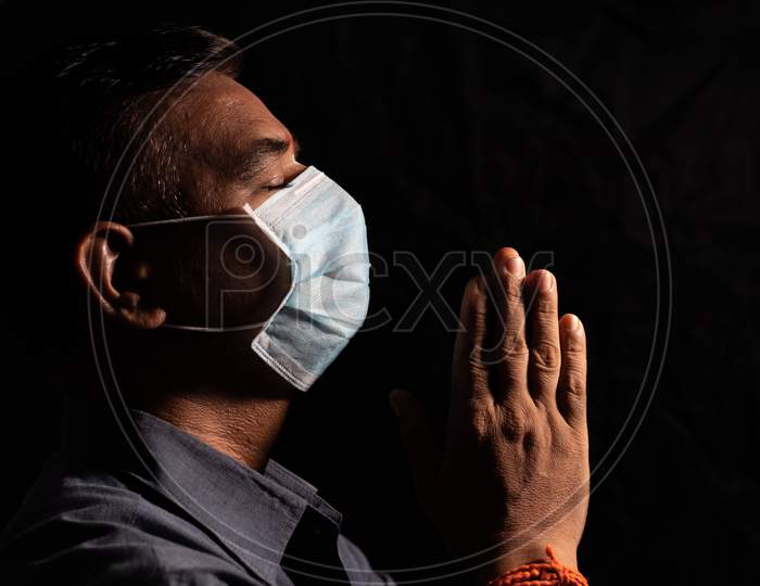 Man With Medical Mask Praying To God By Closing Eyes In Dark Room To Protect Or Save From Covid-19 Or Coronavirus Crisis - Spirituality And Surrender Concept.