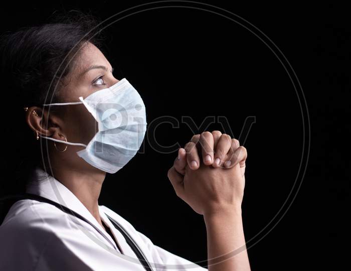 Profile View Of Young Woman Doctor In Medical Mask Praying To God On Black Background Looking Up - Concept Of Hope And Fight To End Coronavirus Or Covid-19 Crisis.