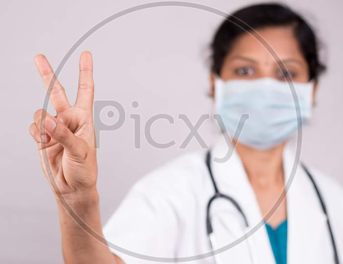 Woman Doctor With Medical Mask In Uniform Showing V Gesture On Isolated Background - Concept Of Victory Salute Or Peace Gesture By Doctor During Covid-19 Or Coronavirus Pandemic.