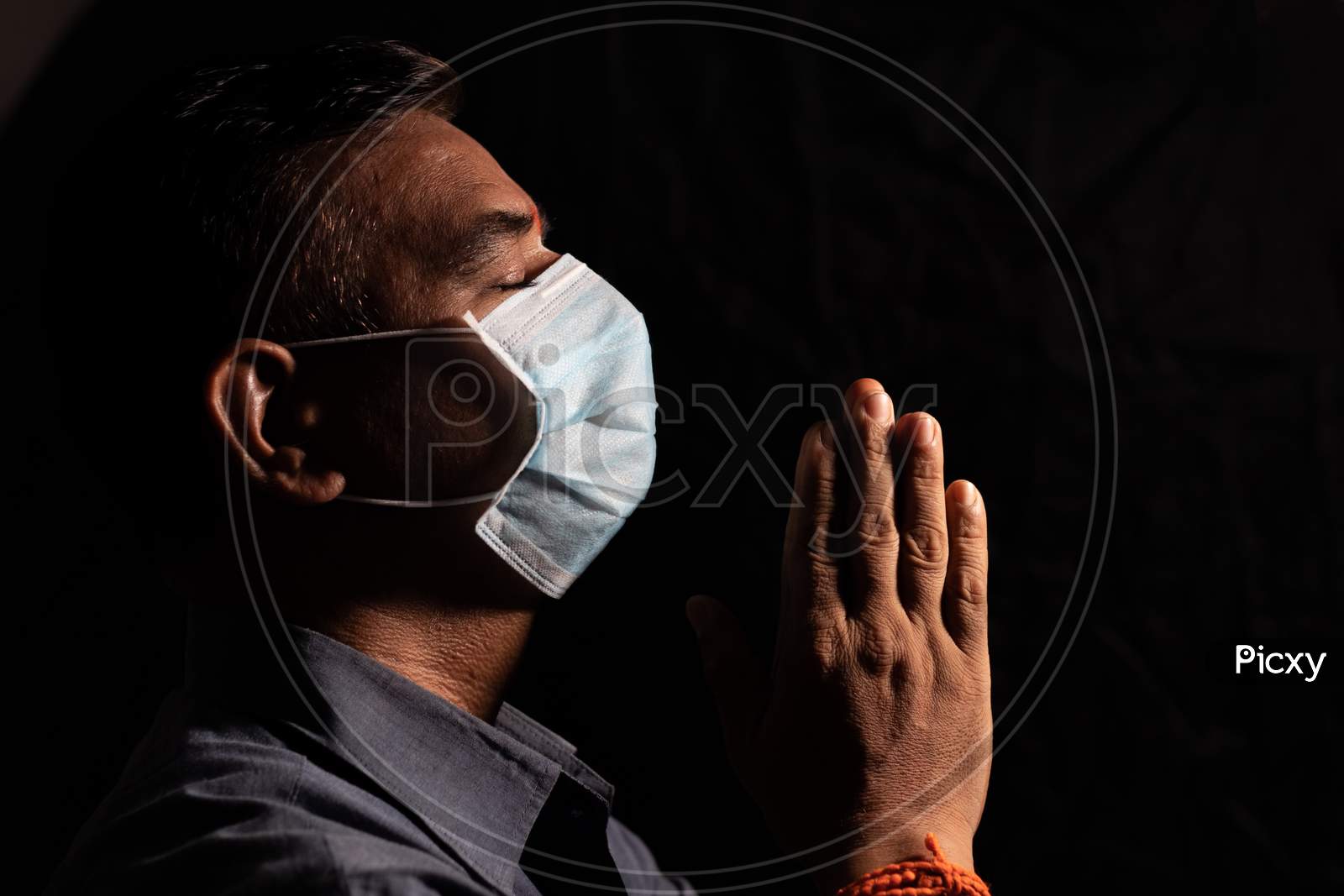 Man With Medical Mask Praying To God By Closing Eyes In Dark Room To Protect Or Save From Covid-19 Or Coronavirus Crisis - Spirituality And Surrender Concept.