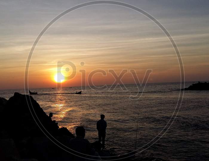 A Silhouette Landscape Image Of People Watching Sunset On A Beach With Fishing Boats In Kerala, India.