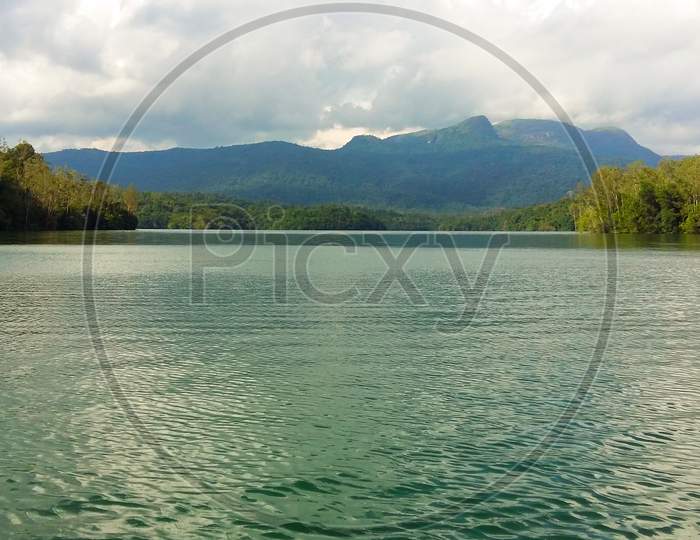 A Landscape Image Of Lake With Greenish Water And Green Trees And Mountains On The Background With Cloudy Sky. Greenish Tint. Neyyar Wildlife Sanctuary.