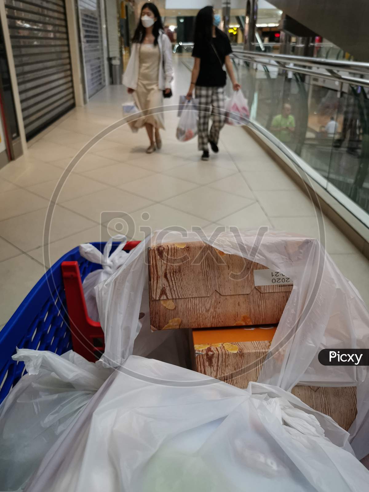 Corona virus safe shopping, the photo shows a shopping cart in the foreground and two walking women wearing medical masks and carrying shopping bags