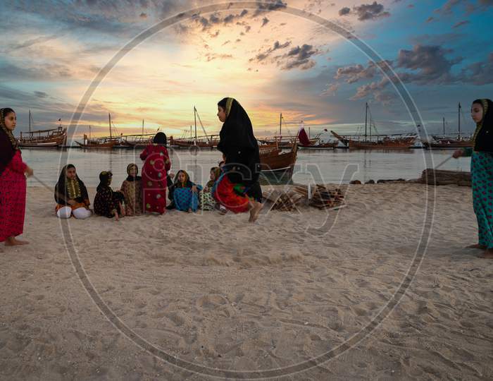 Qatari girls in traditional costume  jumping rope in Katara beach at sunset with dhows in the water and clouds in sky in background