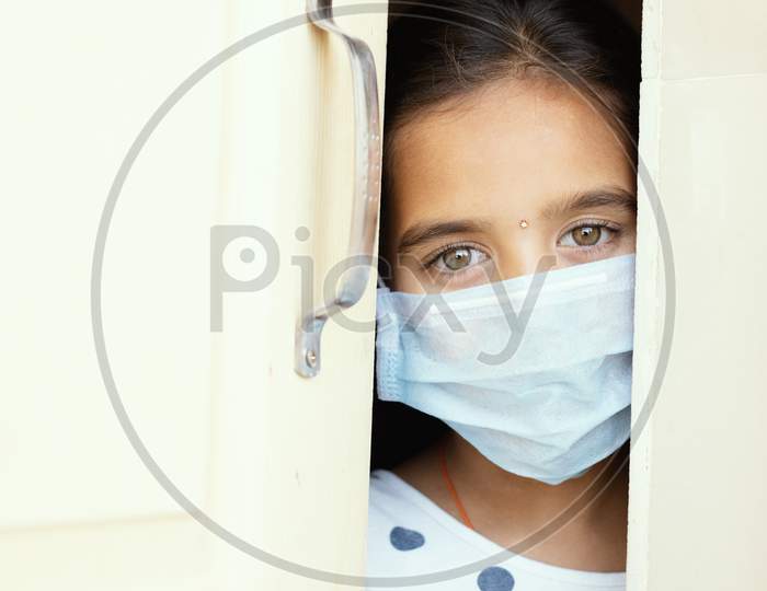 Young Sad Girl With Medical Mask Wearing Sneaking Out Through The Home Door - Concept Of Home Quarantine Due To Covid-19 Or Coronavirus Outbreak.