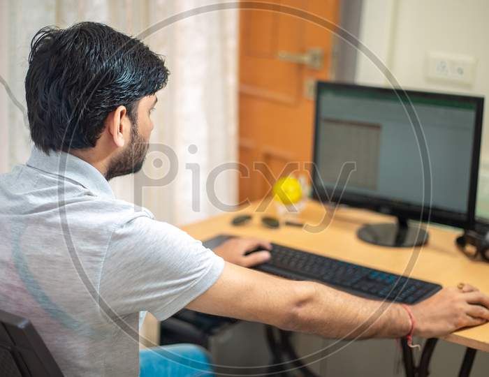 Close Up Rear View Of Indian Man Sitting At A Table At Home Working On A Computer. Freelancer Working From Home.Young Male Student Typing On Computer Wearing Casual Cloths.