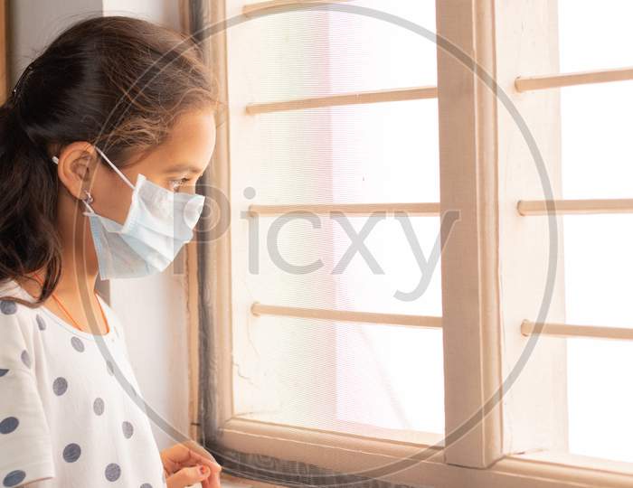 Sad Young Girl Seeing Through The Window During Home Isolation Watching Out - Coronavirus Or Covid-19 Quarantine Concept.