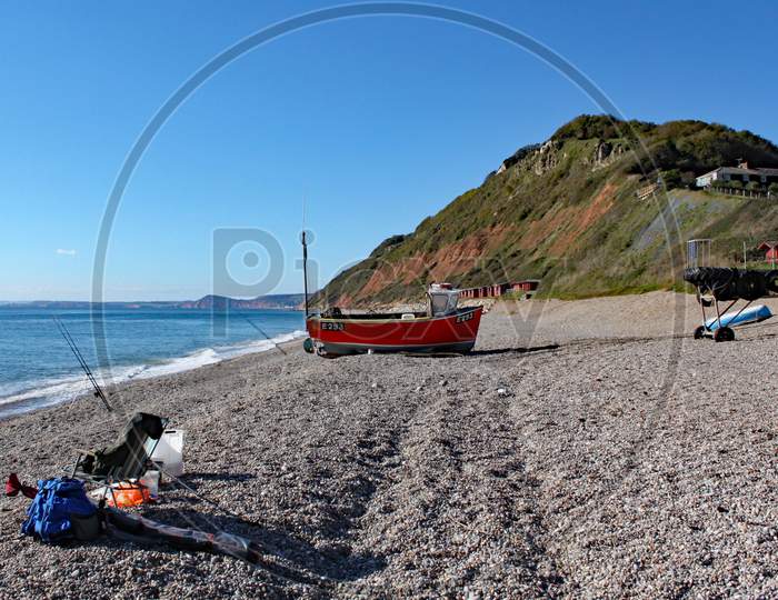 An Old Boat On The Beach At Branscombe In Devon, England. Fishermen'S Equipment Stands In The Foreground.