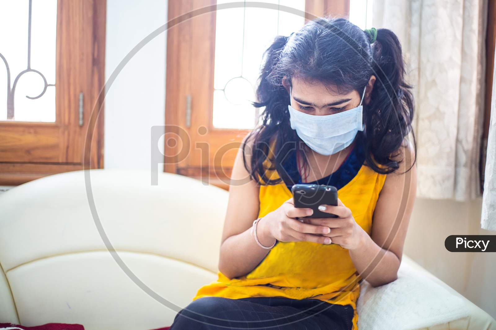 Young Girl Quarantined At Home During World Pandemic Of Coronavirus Covid-19 Prevention. Stay At Home, Stay Safe. Girl With Protective Medical Mask Using Smatphone. Isolation, Lock Down Situation.