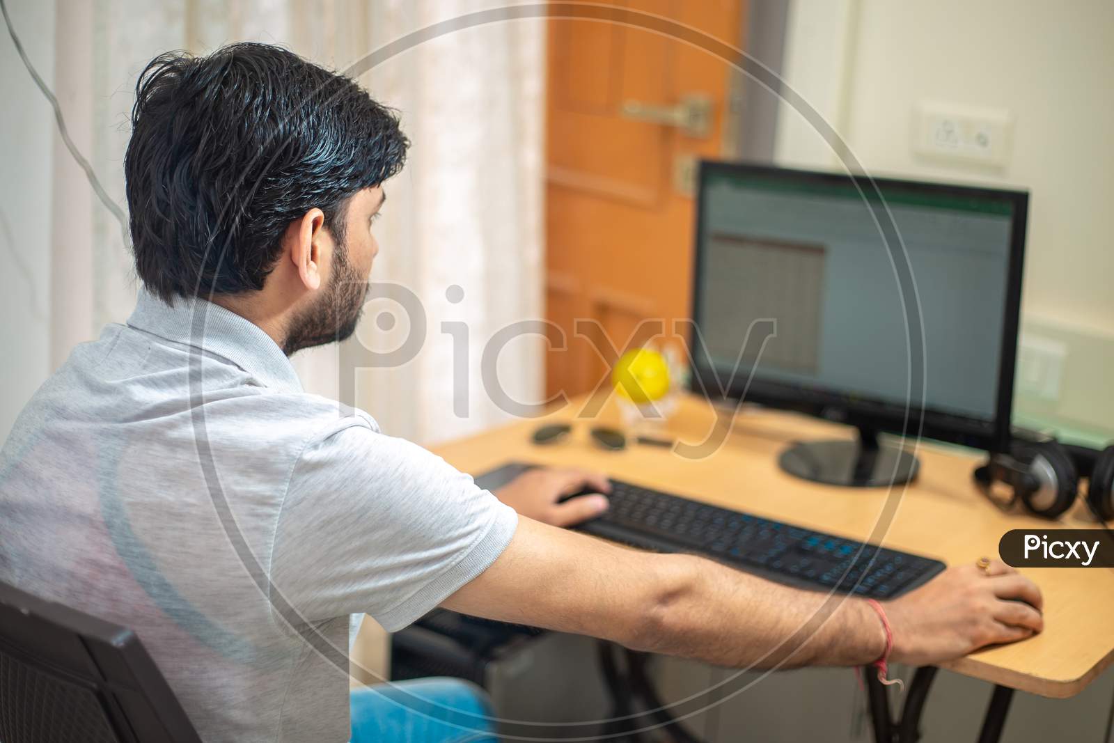 Close Up Rear View Of Indian Man Sitting At A Table At Home Working On A Computer. Freelancer Working From Home.Young Male Student Typing On Computer Wearing Casual Cloths.