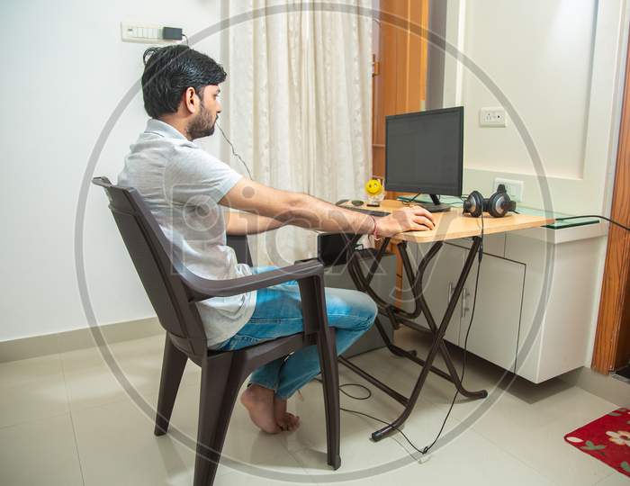 Young Indian Man Sitting At A Table At Home Working On A Computer. Side View.Freelancer Working From Home.Young Male Student Typing On Computer Wearing Casual Cloths.