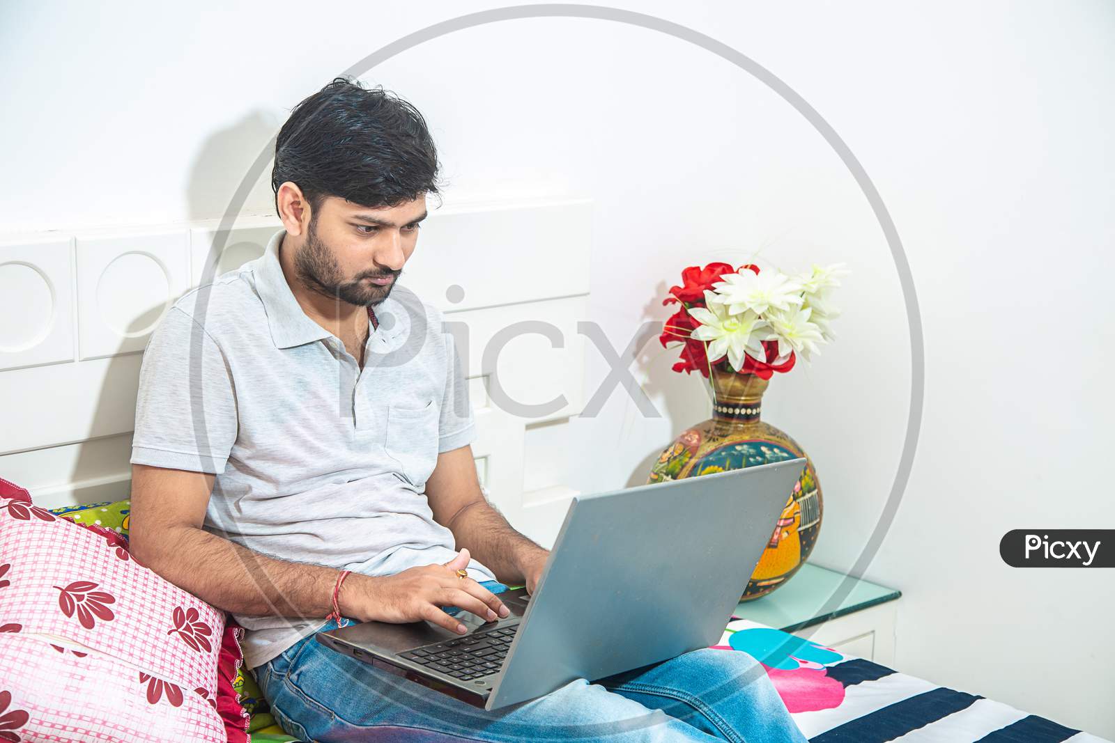 Close Up Of Young Indian Man Busy Working On His Laptop Doing Office Work While Relaxing On Bed In Bedroom, Freelancer Working From Home. Young Male Student Typing On Computer Wearing Casual Cloths.