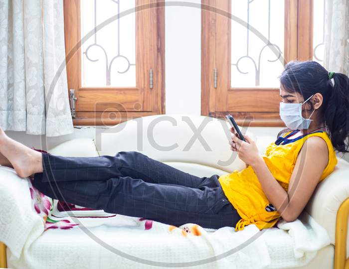 Young Girl Quarantined At Home During World Pandemic Of Coronavirus Covid-19 Prevention. Stay At Home, Stay Safe. Girl With Protective Medical Mask Using Smatphone. Isolation, Lock Down Situation.