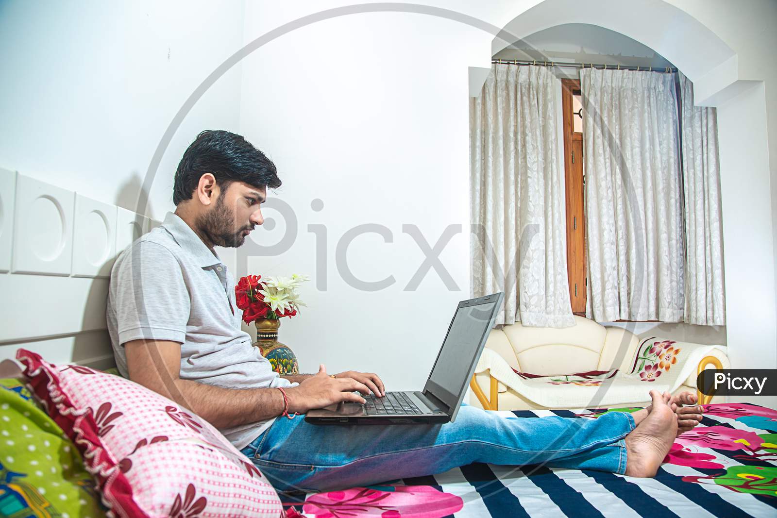 Young Indian Man Busy Working On His Computer Doing Office Work While Relaxing On Bed In Bedroom, Freelancer Working From Home. Young Male Quarantine Himself Coronavirus Outbreak Lock Down Concept.