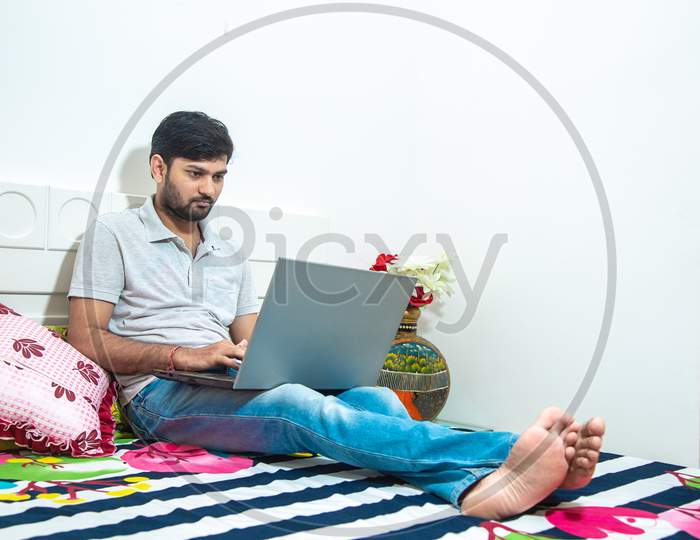 Young Indian Man Busy Working On His Laptop Doing Office Work While Relaxing On Bed In Bedroom, Freelancer Working From Home. Young Male Student Typing On Computer Wearing Casual Cloths.