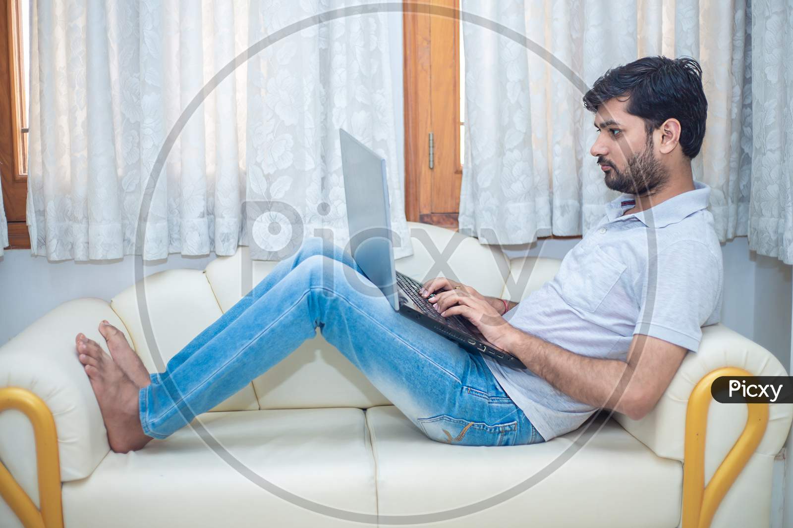 Young Indian Man Working On His Laptop While Relaxing On Sofa In Living Room, Freelancer Working From Home. Young Male Student Typing On Computer Wearing Casual Cloths. Home Office.