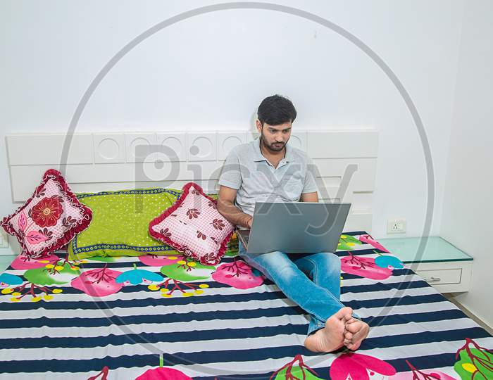 Young Indian Man Busy Working On His Laptop Doing Office Work While Relaxing On Bed In Bedroom, Freelancer Working From Home. Young Male Student Typing On Computer Wearing Casual Cloths.