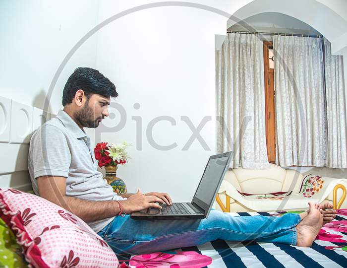 Young Indian Man Busy Working On His Computer Doing Office Work While Relaxing On Bed In Bedroom, Freelancer Working From Home. Young Male Quarantine Himself Coronavirus Outbreak Lock Down Concept.