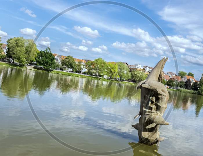 A Dolphin Sculpture In A Small Lake Of A Public Park Below A Cloudy Summer Sky