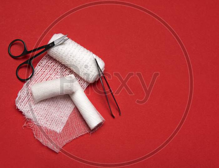 White Medical Bandages Rolls With Scissors And Tweezers In A Pharmacy Kit On A Red Background With Space For Copy