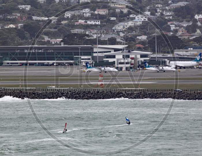 Two Wind Surfers On Lyall Bay In Wellington New Zealand On A Grey Stormy Day. The Airport Can Be Seen In The Background.