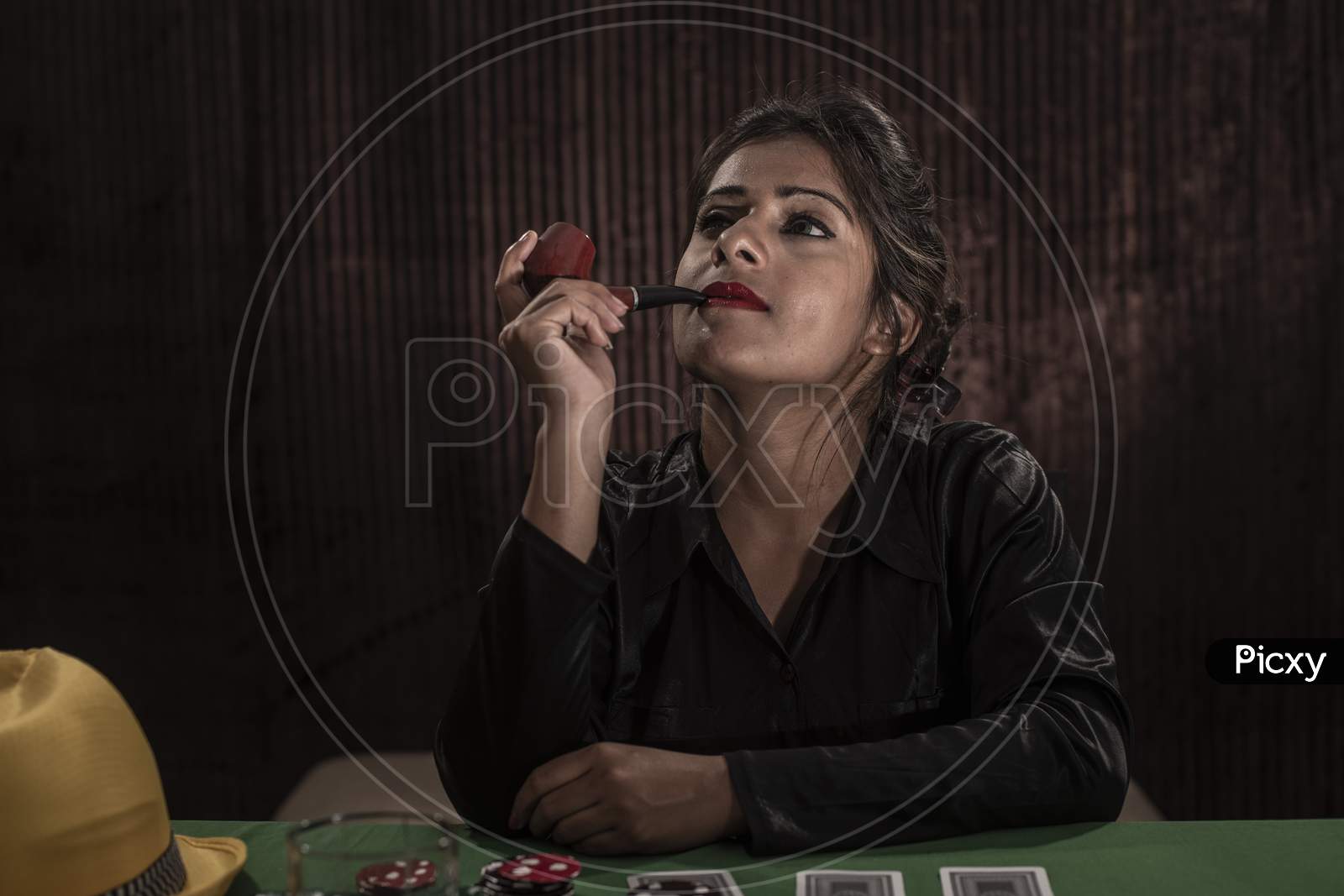Young Indian Bengali brunette woman in western dress with pipe playing cards on a casino poker table in brown textured copy space studio background. Indian lifestyle and fashion.