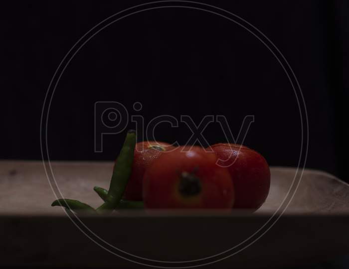 Fresh red tomatoes and green chilies are kept on a wooden tray in a dark copy space background