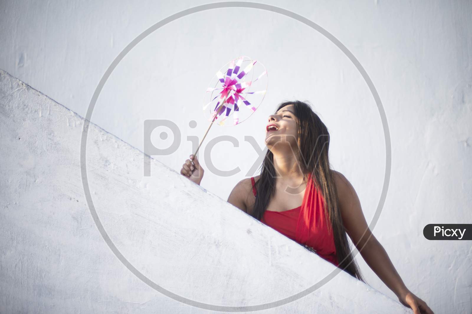 An young and attractive Indian brunette woman in red western dress enjoying with a pinwheel on the rooftop in a morning. Indian lifestyle and quarantine.