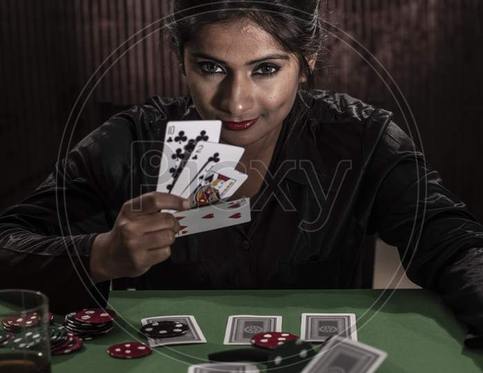 Young Indian Bengali brunette woman in western dress with pipe playing cards on a casino poker table in brown textured copy space studio background. Indian lifestyle and fashion.