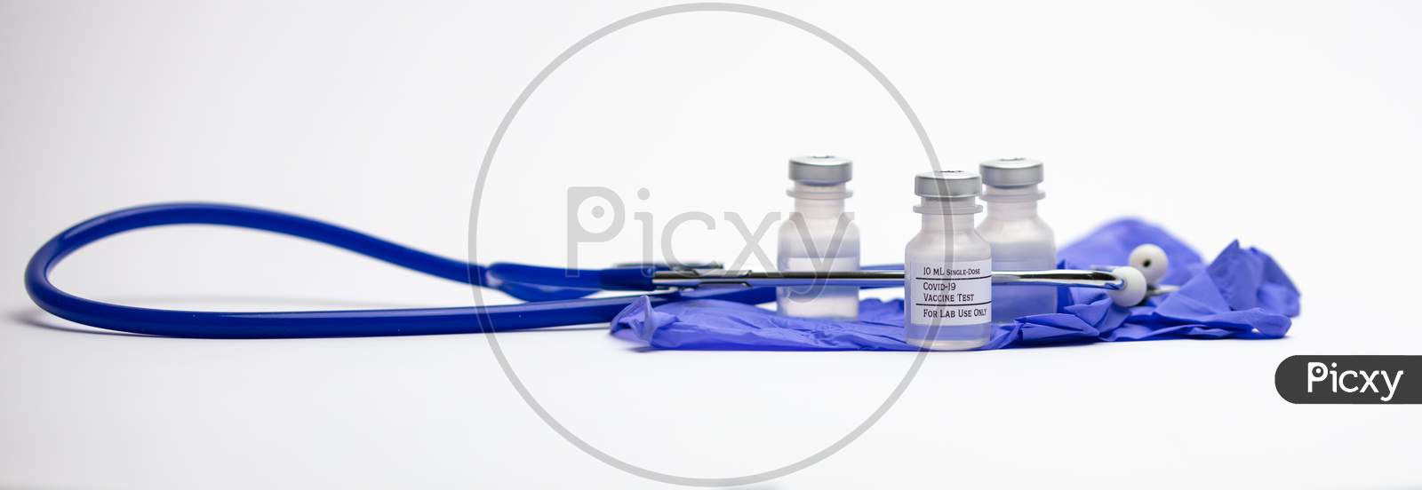 Three Covid-19 Test Vaccine Vials On Top Of Blue Medical Gloves And Surrounded By A Blue Stethoscope.