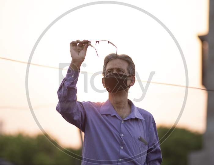 portrait of an Indian old man with corona preventive mask looking at his spectacles on a rooftop during sunset in home isolation.Indian lifestyle, disease and home quarantine.